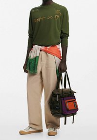 Camicia a maniche lunghe verde oliva con testo stampato, pantaloni khaki a gamba larga, sciarpa colorata avvolta e zaino verde scuro con molte tasche.
