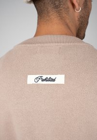 Vue de dos d'une personne portant un pull en maille beige avec une étiquette blanche et un texte en écriture cursive noire indiquant "Prohibited" sur le haut du dos.