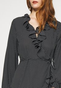 Svart prickig klänning med volangkrage och långa ärmar. Tyget är glatt, med en figursydd midja och subtilt knytband på sidan.