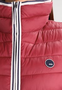 Col montant en tissu matelassé rouge avec un biais de fermeture éclair rayé blanc et marine, et un patch rond de logo marine sur la poitrine.