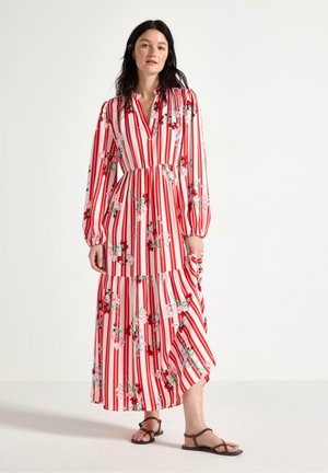 Kvinde med mørkt hår iført lang, rød- og hvidstribet kjole med blomsterprint og lange ærmer, stående og holdende i en del af skørtet.