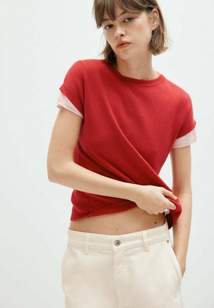 Jeune femme aux cheveux bruns courts portant un haut rouge à manches courtes par-dessus une chemise rose et un pantalon blanc, levant son haut pour révéler son ventre.