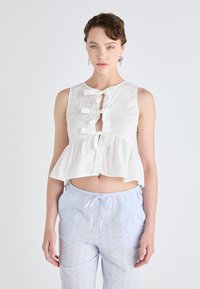 Crop top bianco in cotone con design peplum, caratterizzato da tre fiocchi e un'apertura a chiave. Abbinato a pantaloni in cotone azzurro chiaro con coulisse.