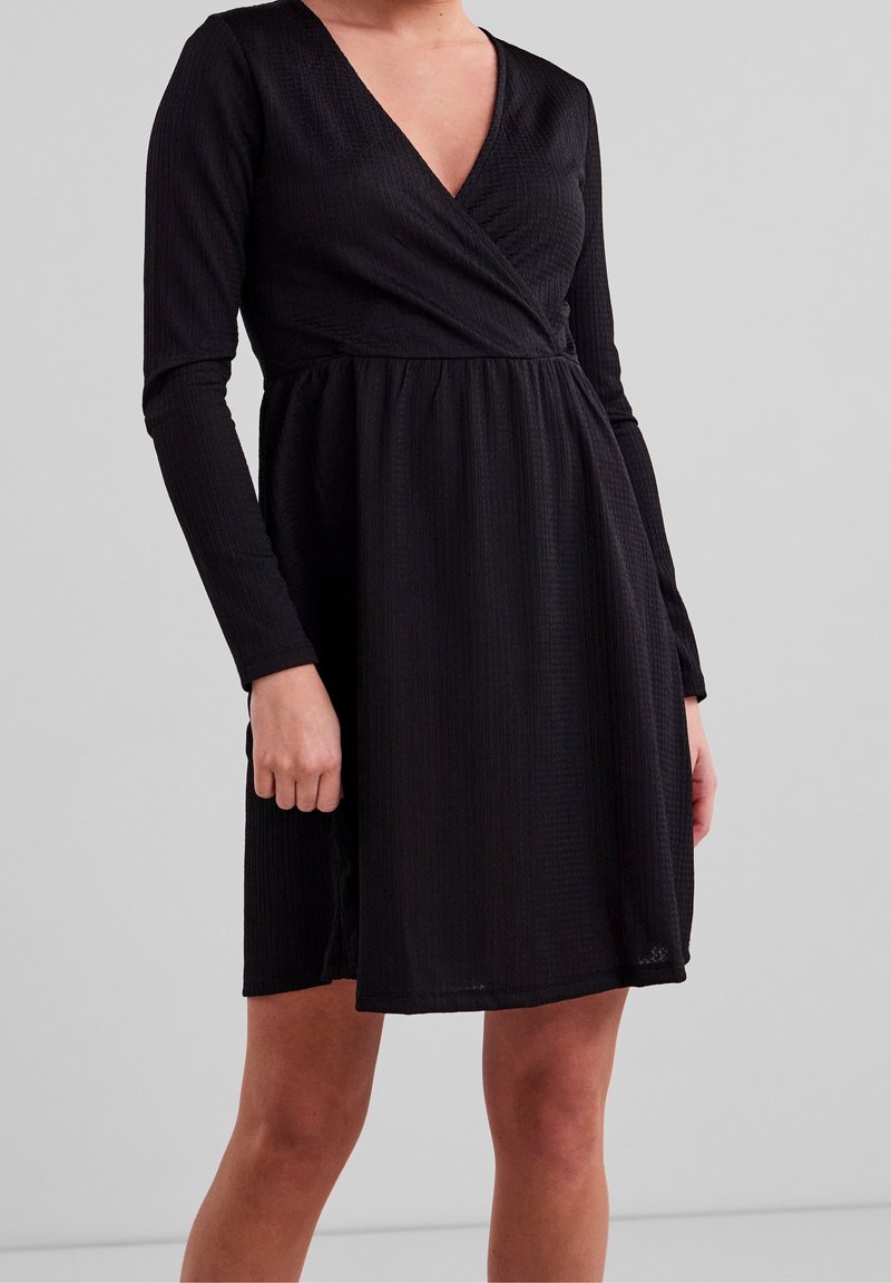 Robe noire à manches longues avec un décolleté en V profond, en tissu texturé, corsage ajusté et jupe évasée, présentant un design plissé subtil.