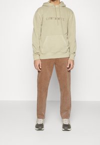 Huppari beige värviga, millel on ees tasku ja tikandiga logo. Pruunid kašmiirkangast püksid, lahtine lõige ja hallid tennisetossud mustade aktsentidega.
