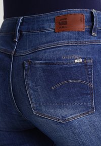 Blå denimjeans med en brun läderlapp som har "RAW"-logotypen, femficksdesign, synliga sömmar och tan accenter.