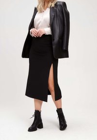 Femme portant un chemisier blanc, un blazer en cuir noir posé sur les épaules, une jupe midi noire avec une fente latérale, et des bottines noires à lacets.