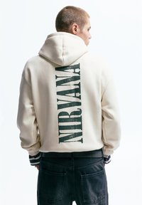 Crèmekleurige hoodie met een grote zwarte "NIRVANA" print op de achterkant. Voorzien van een capuchon met trekkoord en geribbelde manchetten met zwart-witte strepen.