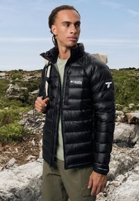 Sort isoleret jakke med horisontal quiltning, der har en høj krave og Columbia Titanium-logo. Båret over en lysegrøn skjorte.