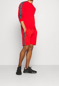 Ensemble de sweat-shirt athlétique rouge et short, avec des bandes latérales noires ornées de logos. Coupe confortable, confectionné dans un tissu doux, accompagné de baskets noires.