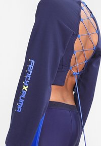 Granatowa bluzka z długim rękawem z wycięciem na plecach, ozdobiona niebieskim sznurowaniem i napisem na mankietach "Fenty x Puma". Materiał wydaje się elastyczny i gładki.