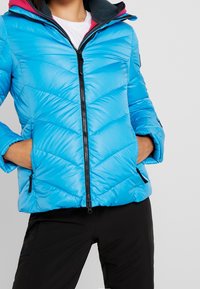 Veste en duvet bleue avec un design matelassé, dotée d'une fermeture éclair noire et de deux poches latérales. Doublée avec des accents roses sur la capuche.