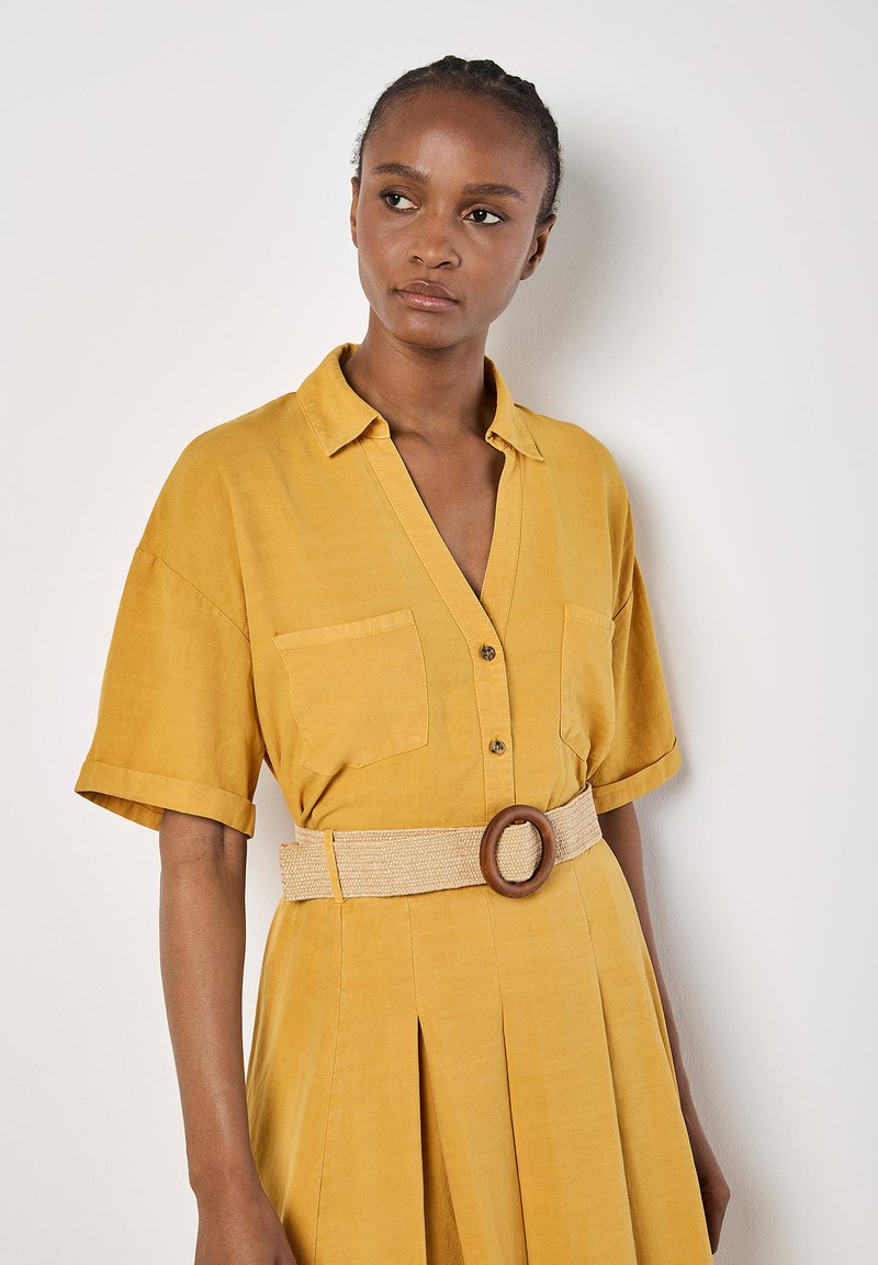 Senfgelbes Kleid mit kurzen Ärmeln, Knopfleiste vorne, zwei Brusttaschen und einem gewebten Gürtel mit einer Holzring-Schnalle.