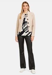 Beżowa krótka kurtka z zapięciem na guziki, z dwoma przednimi kieszeniami, noszona na czarno-białą graficzną bluzkę, w zestawieniu z czarnymi spodniami w stylu dzwonów i białymi trampkami.