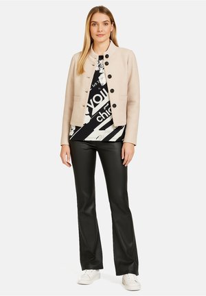 Beżowa krótka kurtka z zapięciem na guziki, z dwoma przednimi kieszeniami, noszona na czarno-białą graficzną bluzkę, w zestawieniu z czarnymi spodniami w stylu dzwonów i białymi trampkami.