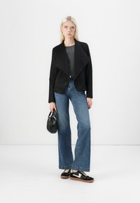 Svart, drapert jakke over en grå topp, parret med blå flagrende jeans og svarte joggesko. Tilbehør med en liten svart håndveske.