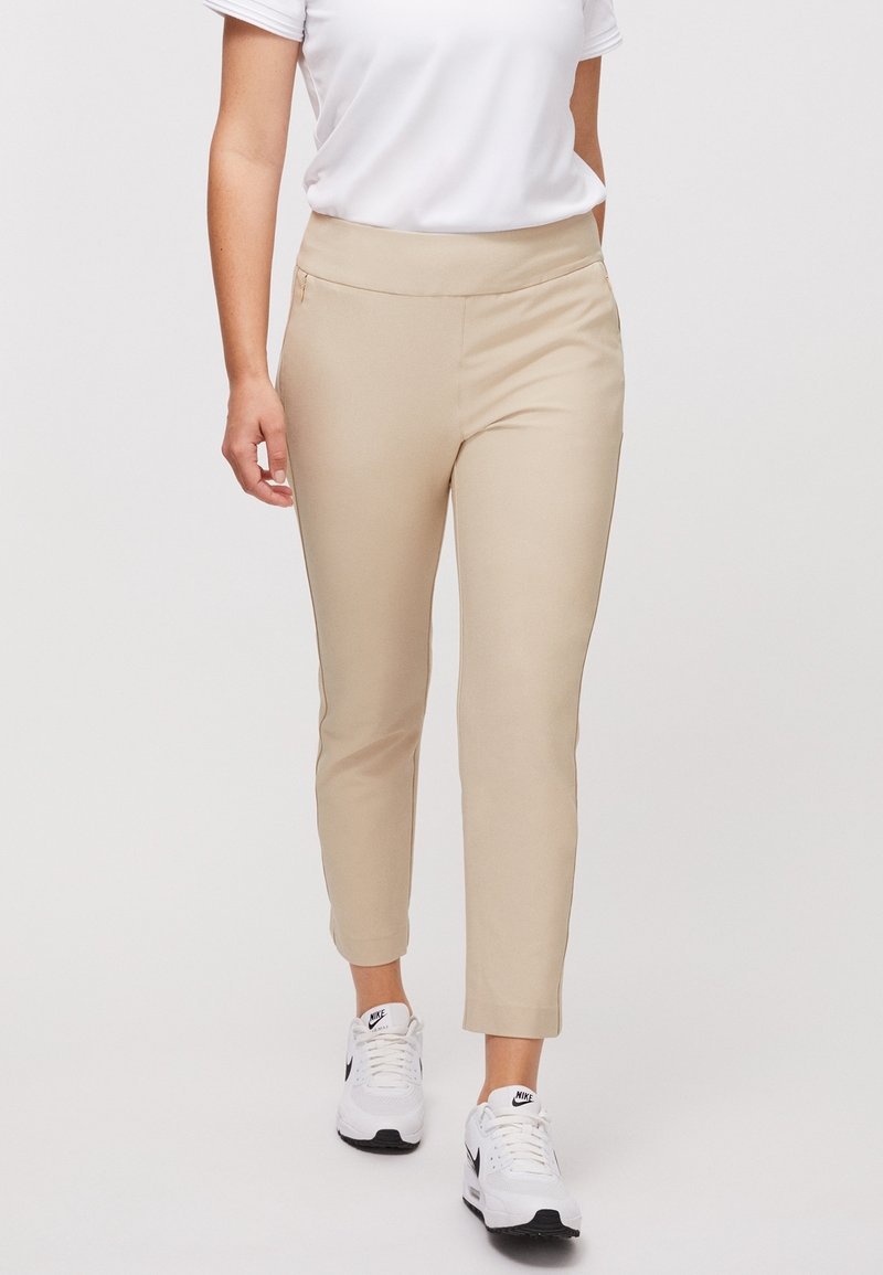 Beige elastické kalhoty ke kotníkům s hladkým pasem a bočními kapsami, spárované s bílými teniskami.