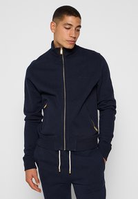 Homme portant une veste zippée bleu marine avec une fermeture éclair dorée et un pantalon bleu marine assorti, se tenant les mains dans les poches, regardant vers le bas et sur le côté.