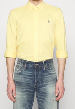 Homme portant une chemise jaune clair boutonnée avec les manches retroussées et un jean bleu délavé, debout devant un fond uni.