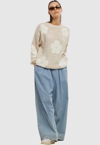 Beiger Pullover mit großen weißen Blumenmustern, kombiniert mit lockeren hellblauen Jeanshosen. Modell mit dunklen Sonnenbrillen und Turnschuhen.