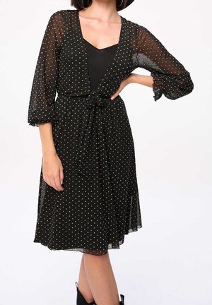 Kvinde iført en sort kjole til knæet med gennemsigtige ærmer med polkaprikker og bindebånd i taljen, kombineret med sorte ankelstøvler.