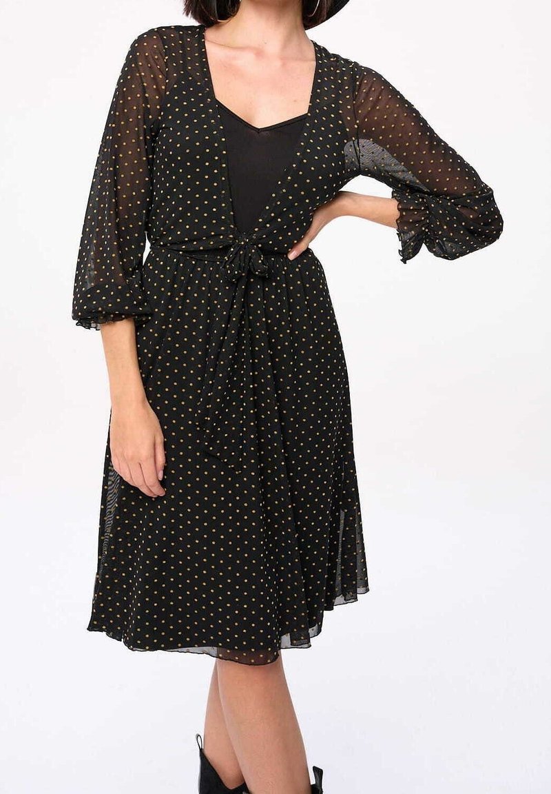 Femme portant une robe noire longueur genou avec des manches en tulle à pois et une taille nouée, associée à des bottines noires.