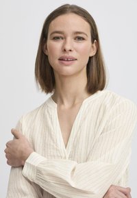 Blusa leggera color crema con righe verticali in un tessuto morbido, con scollo a V, maniche lunghe e dettagli con bottoni sulla parte anteriore.