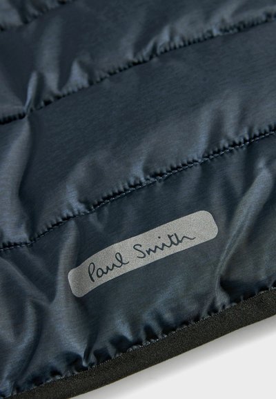 Mīkstināta tumši zila auduma ar spīdīgu faktūru, ar sudraba etiķeti, kurā ir rakstīts "Paul Smith" stilizētā rokrakstā. Šuves ir redzamas gar šuvēm.