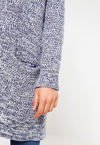Pletený svetr v námořnické modré a bílé barvě, se strukturovaným vzorem, dlouhými rukávy a přední kapsou. Tkanina vypadá měkce a teple.