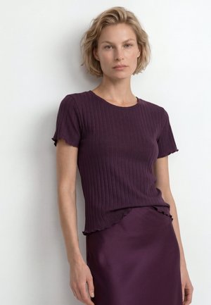 Mujer rubia con el pelo corto que lleva una camiseta morada de manga corta con costillas y una falda a juego sobre un fondo blanco liso.