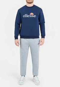 Marineblauwe sweatshirt met "ellesse" logo, gecombineerd met lichtgrijze taps toelopende joggingbroek. Witte sneakers maken de sportieve outfit compleet.