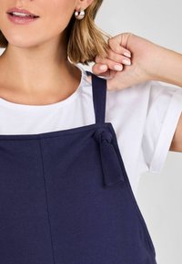 Marineblaues Schürze mit einem Knotendetail am Schulterträger, getragen über einem weißen T-Shirt. Glattes Material und strukturierte Form sichtbar.
