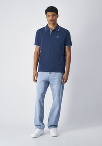 Granatowa koszulka polo z białymi wykończeniami i logo, w zestawieniu z jasnoniebieskimi prostymi dżinsami i białymi trampkami. Gładki bawełniany materiał.