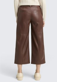 Braune Lederimitat-Hose mit weitem Bein und hoher Taille, ausgestattet mit zwei Gesäßtaschen und einer glatten Textur, kombiniert mit cremefarbenen Plateauschuhen.
