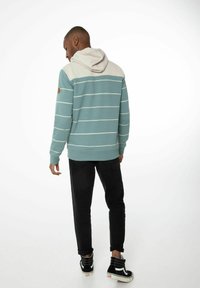 Groene hoodie met witte strepen, met een lichtgrijze capuchon, geribde manchetten en een logo patch op de linkerarm. Zwarte broek en sneakers.