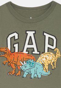 Maslinasta košulja s bijelim natpisom "GAP" na kojem se nalaze crveni T-Rex, plavi triceratops i žuti parasaurolophus ispod slova.