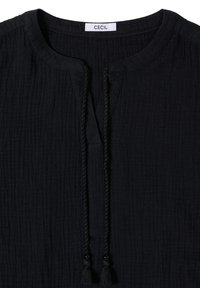 Blouse noire texturée avec encolure ronde et liens tressés à pompons sur le devant, étiquetée "CECIL" à l'intérieur du col.