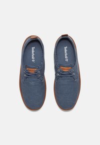 Zapatillas de lona azules con suelas de goma marrón, punta redonda y diseño de cordones. Presenta un logo en la plantilla y un acento de cuero en la lengüeta.