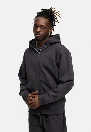 Dark gray zip-up hoodie made of soft fabric with a front pocket and a drawstring hood. Model presents a relaxed fit with ribbed cuffs.