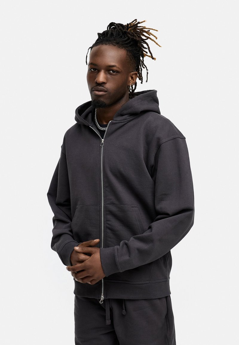 Dunkelgrau zip-up Kapuzenpullover aus weichem Material mit einer Fronttasche und einer Kordelzugkapuze. Das Modell zeigt eine lässige Passform mit gerippten Bündchen.