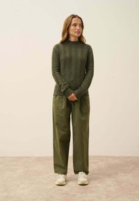 Olive zielony sweter robiony na drutach z teksturowanymi paskami, w połączeniu z luźnymi olive zielonymi spodniami i jasnymi trampkami, stojący na neutralnym tle.