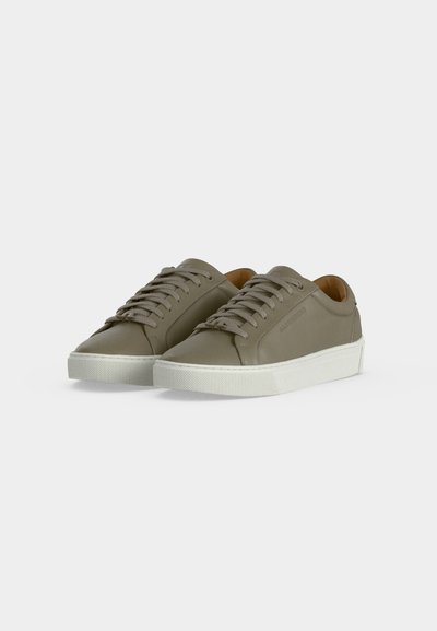Paire de baskets basses vert olive avec semelles en caoutchouc blanches et lacets assortis sur fond gris clair.