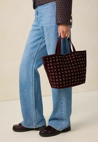 Bordeaux suède tote bag met witte oogjespatronen, vastgehouden door een persoon in lichtblauwe uitlopende jeans en donkere loafers met vooraccenten.