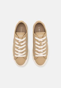 Beige syntetiska sneakers med vita snörning, runt tå och gummisulor. Har varumärkeslogotyp på sidan och en texturerad innerfoder.
