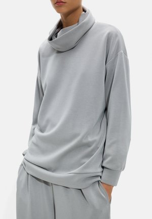 Osoba w jasnoszarej bluzce z długim rękawem, luźnym kołnierzem typu komin i dopasowanych, wygodnych spodniach, ręka w kieszeni na białym tle.