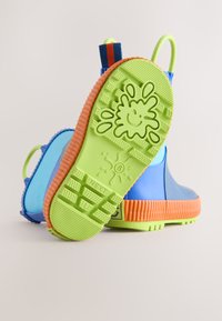 Bottes de pluie enfant bleues et orange avec semelles vertes vives, présentant un motif éclaboussé souriant et un motif de semelle texturé.