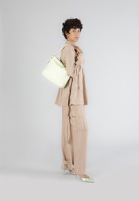 Světle zelená kožená kabela typu bucket s pletenou rukojetí, nošená přes béžový lněný outfit s velkými kapsami a přizpůsobeným střihem, doplněná mátovými podpatky.