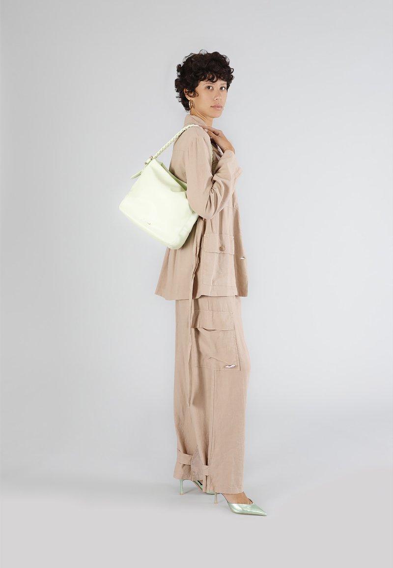 Světle zelená kožená kabela typu bucket s pletenou rukojetí, nošená přes béžový lněný outfit s velkými kapsami a přizpůsobeným střihem, doplněná mátovými podpatky.