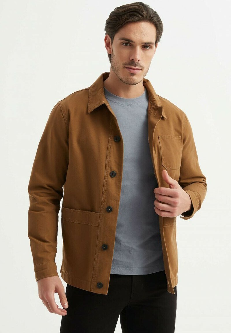 Veste utilitaire marron avec des boutons noirs, dotée d'une poche poitrine et de poches latérales, portée sur un T-shirt gris clair, avec un col classique.