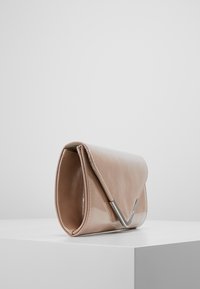 Béžová lesklá kožená kabelka s trojúhelníkovým stříbrným kovovým uzávěrem odpočívající na bílém podstavci na šedém pozadí.